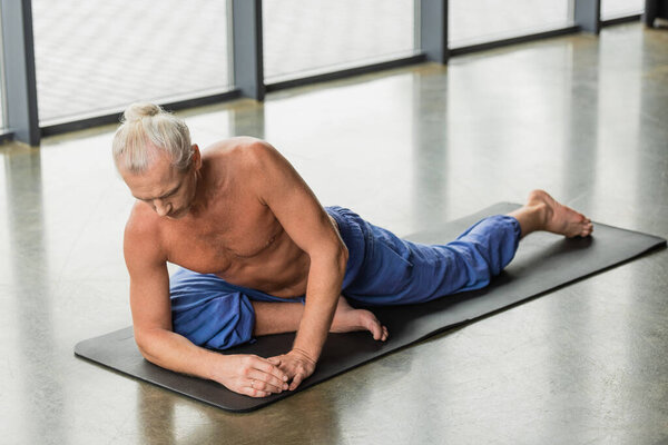 grey haired man doing half pigeon pose on mat in yoga studio 