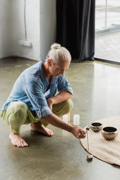 stock image grey haired man burning incense stick near Tibetan singing bowls and candles in yoga studio 