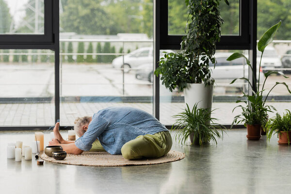 grey haired man stretching back near Tibetan singing bowls and candles in yoga studio 