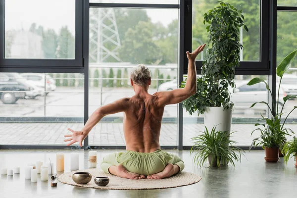 stock image back view of shirtless man gesturing while sitting in thunderbolt yoga pose near candles and Tibetan singing bowls 