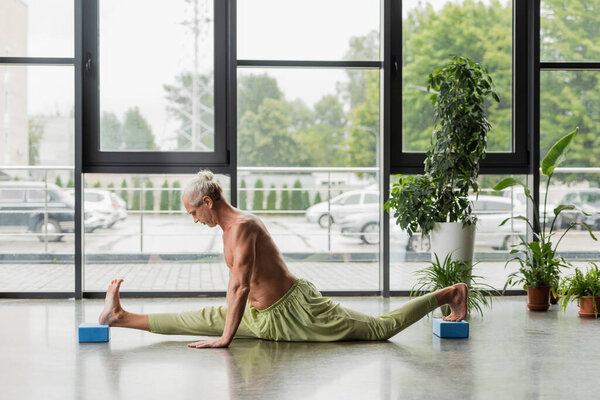 side view of shirtless and grey haired man doing split on yoga foam blocks 