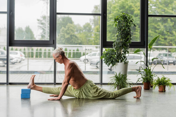 side view of shirtless and grey haired man doing split on blue yoga foam blocks in studio 