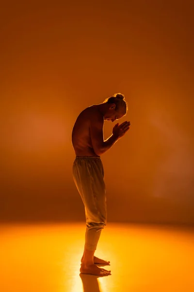 full length of shirtless man in pants practicing yoga with praying hands on orange background