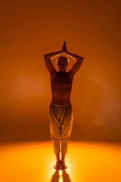 full length of shirtless man in pants standing in warrior pose on orange background