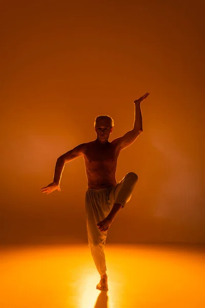 full length of shirtless man in pants practicing yoga on orange background
