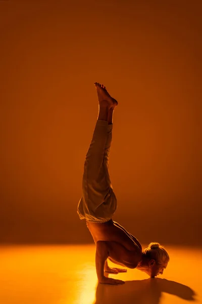 full length of shirtless man in pants doing chin stand yoga pose on brown