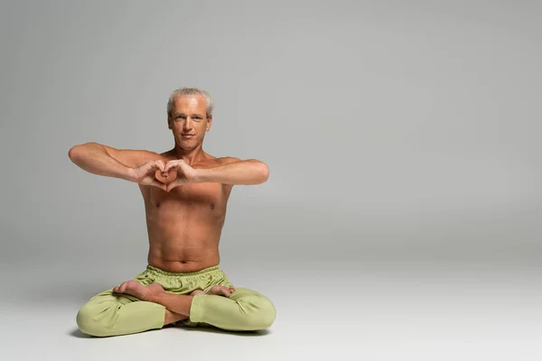 Stock image shirtless man in green pants sitting in lotus pose and showing heart sign on grey background 