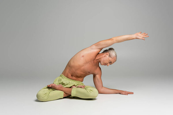 shirtless man in green pants sitting in lotus pose and doing side bend on grey background 