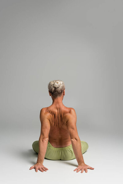 back view of shirtless man in green pants sitting in lotus pose on grey background 