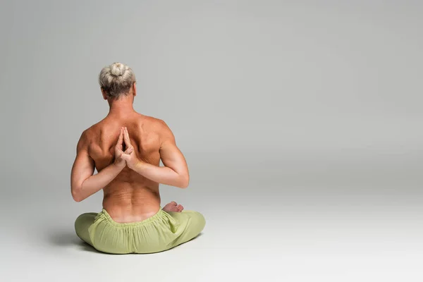 stock image back view of shirtless man in green pants sitting in lotus pose and doing anjali mudra behind back on grey background 