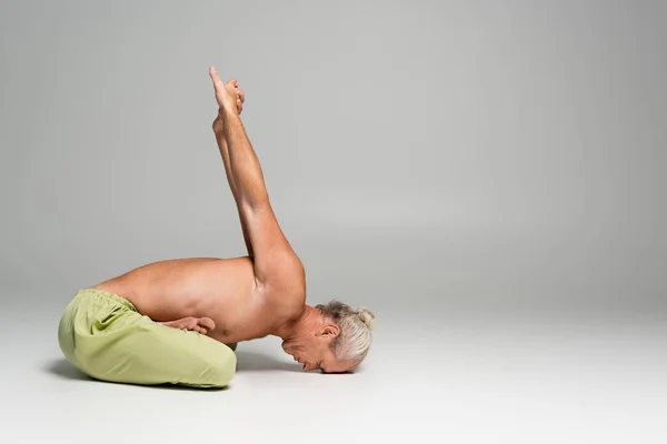 side view of shirtless man in pants sitting in lotus pose and stretching back on grey background
