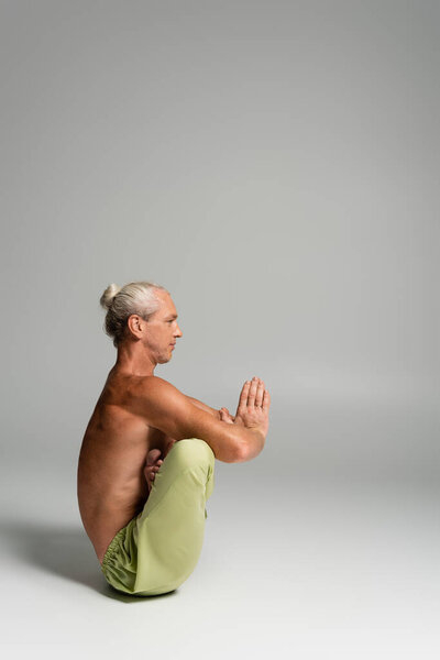 боковой вид мужчины без рубашки в штанах, сидящего в позе эмбриона матки и делающего Анджали мудру на сером 