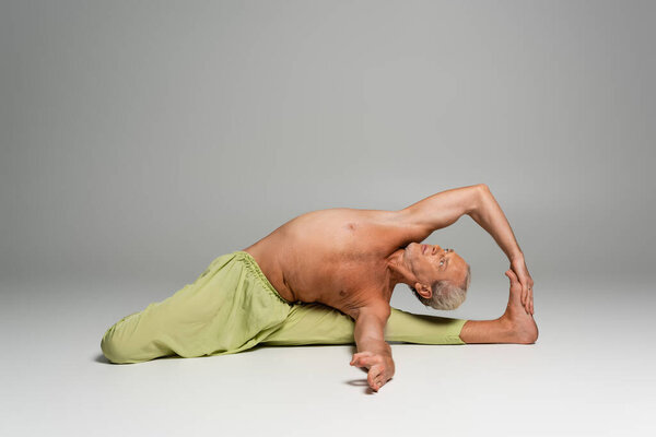 shirtless man in pants doing seated gate yoga pose on grey background 