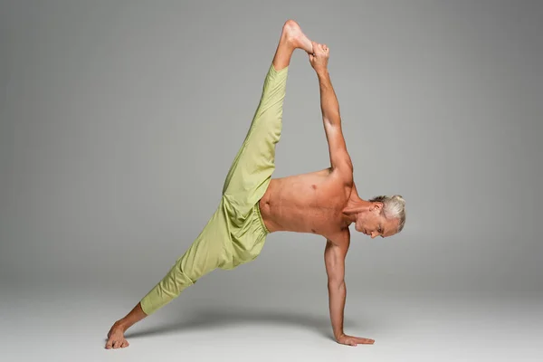 full length of barefoot man in pants doing side plank yoga on grey background