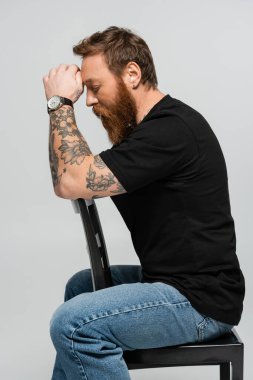 side view of tired bearded man in black t-shirt and wristwatch sitting with closed eyes and hands near head isolated on grey