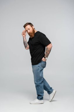 full length of thoughtful bearded man touching forehead and holding hand in back pocket of jeans on grey background