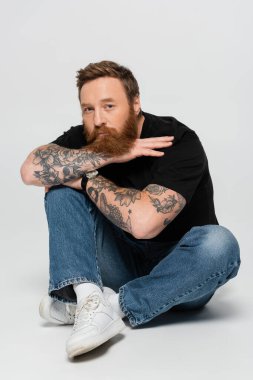 stylish tattooed man in blue jeans and white sneakers sitting with crossed legs and looking at camera on grey background