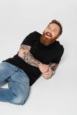 joyful bearded and tattooed man in jeans and black t-shirt laughing while lying on grey background