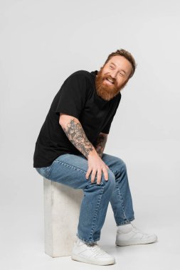 cheerful tattooed man in black t-shirt and jeans sitting on construction block and looking away on grey background