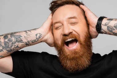 portrait of overjoyed bearded man touching head and laughing with closed eyes isolated on grey