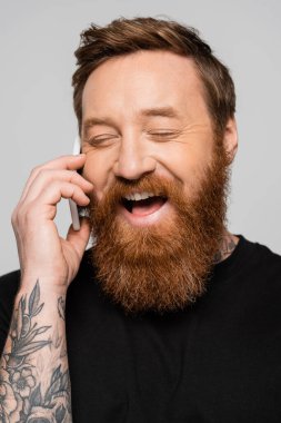 excited bearded man with closed eyes talking on mobile phone and laughing isolated on grey