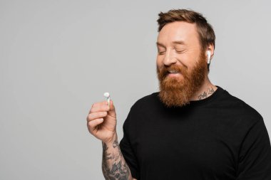 positive bearded man in black t-shirt looking at wireless earphone isolated on grey