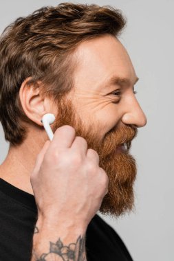 side view of cheerful bearded man holding wireless earphone near ear isolated on grey