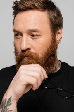 thoughtful bearded man holding eyeglasses and looking away isolated on grey