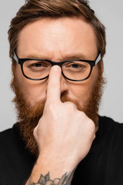 portrait of strict bearded man adjusting eyeglasses and looking at camera isolated on grey