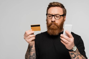 thoughtful and stylish man in eyeglasses holding credit card and mobile phone isolated on grey