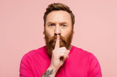frowning bearded man showing hush sign and looking at camera isolated on pink clipart