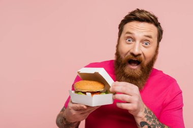 Heyecanlı sakallı adam elinde hamburgerle karton paketle pembe kameraya bakarken