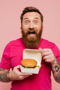 astonished tattooed man in bright t-shirt showing carton pack with delicious hamburger isolated on pink