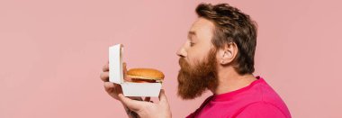side view of bearded man smelling delicious hamburger isolated on pink, banner