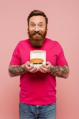 Eflatun tişörtlü mutlu adam pembe üzerine izole edilmiş lezzetli burgerli karton paketi gösteriyor.