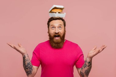 cheerful bearded man in magenta t-shirt posing with delicious burger on head isolated on pink clipart