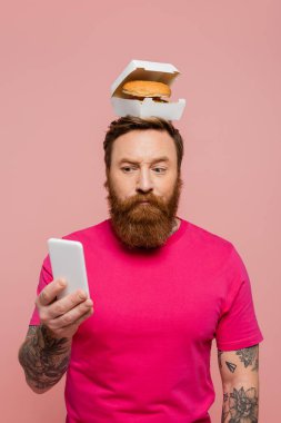 thoughtful bearded man looking at smartphone while standing with tasty burger on head isolated on pink clipart