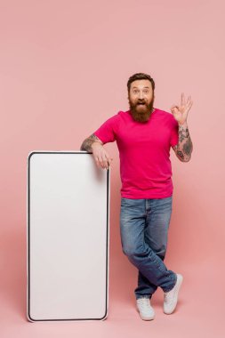 full length of cheerful and stylish man showing okay gesture near big phone template on pink background