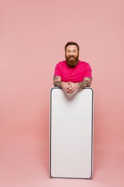 bearded and tattooed man in magenta t-shirt looking at camera near huge phone template on pink background
