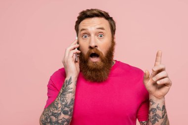 amazed tattooed man talking on mobile phone and showing idea sign isolated on pink