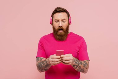 shocked bearded man in wireless headphones using mobile phone isolated on pink