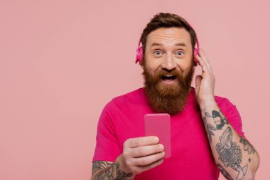 joyful tattooed man with mobile phone touching wireless headphones and looking at camera isolated on pink