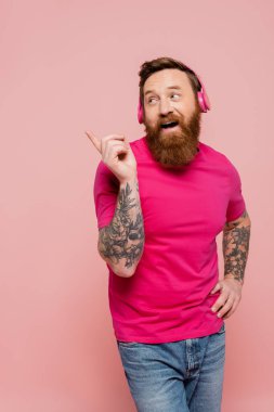 cheerful bearded man in magenta t-shirt and wireless headphones pointing with finger isolated on pink
