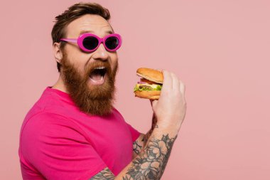 Moda gözlüklü heyecanlı adam ağzını açıyor. Pembeye izole edilmiş lezzetli bir hamburgerin yanında.
