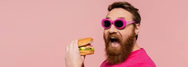 Heyecanlı sakallı şık güneş gözlüklü adam. Hamburgerin yanında ağzını açıyor. Pembe pankartta izole edilmiş.