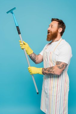 Çizgili önlüklü ve sarı lastik eldivenli gülümseyen adam mavi üzerine izole edilmiş pencere temizleyicisi tutuyor.