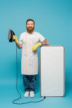 joyful bearded man in apron and rubber gloves posing with iron near big phone mock-up on blue background