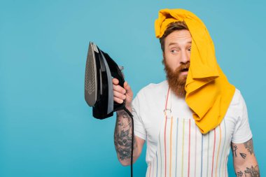 bearded man with yellow garment on head looking at electric iron isolated on blue