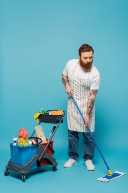 full length of serious bearded man with mop cleaning floor near cart with cleaning supplies on blue background clipart