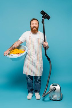 joyful tattooed man in apron holding laundry bowl and vacuum cleaner while looking at camera on blue background